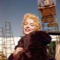 Color photo of Marilyn Monroe taken by Milton H. Greene on the set of the movie Bus Stop in Tahoe in 1956. Marilyn is smiling at the camera wearing a dark fur coat wrapped around her with rustic scaffolding in the background.