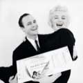 Marilyn Monroe and Marlon Brando are smiling wide as Marlon holds Marilyn with an oversized ticket to the play The Rose Tattoo, a benefit for the Actors Studio. Black and White photo by Milton H Greene in November 1955.